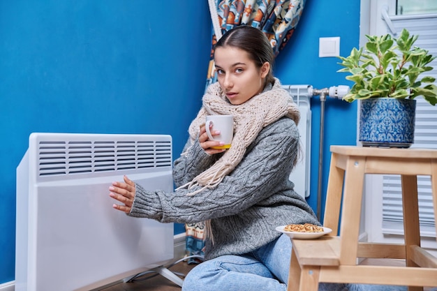 Freddo autunno inverno stagione stile di vita adolescente femmina in maglione caldo sciarpa riscaldamento vicino radiatore riscaldamento elettrico bere bevanda calda con tazza Riscaldamento calore attrezzature vita nella stagione fredda