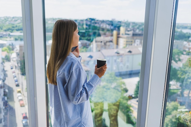 Freelancer donna moderna che parla al telefono stando in piedi davanti a una grande finestra e bevendo caffè Il concetto di una donna moderna di successo Giovane ragazza in un ufficio aperto