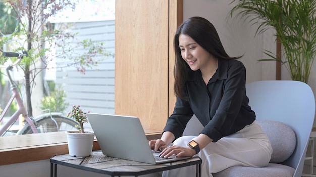 Freelancer femminile abbastanza giovane che si siede sulla poltrona e che utilizza il computer portatile che lavora in linea controllando la posta elettronica