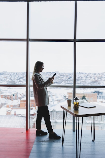 Freelancer femminile concentrato seduto in uno spazio di lavoro moderno con netbook aperto e controllo delle notifiche sul telefono cellulare in abiti casual