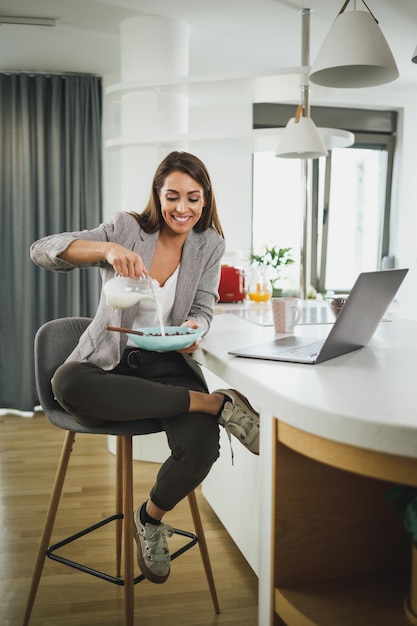 Freelancer femminile sorridente che fa colazione mentre utilizza il laptop a casa.