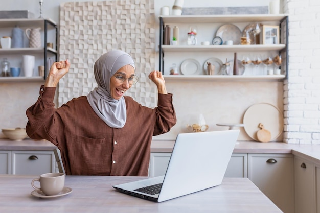 Freelancer in hijab che lavora al laptop da remoto da casa, la donna ha ricevuto buone notizie e buone notizie