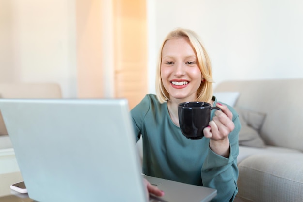 Freelancer positivo della giovane donna che guarda video nel blog sul netbook e che beve un caffè a casa comoda Bella giovane donna d'affari che lavora sul computer portatileFreelancer che si connette a Internet