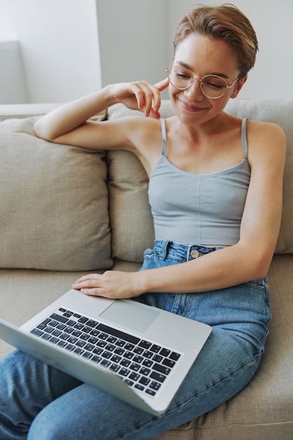 Freelancer ragazza adolescente con laptop seduto sul divano a casa sorridente in abiti e occhiali da casa con stile di vita taglio di capelli corto senza filtri spazio libero copia