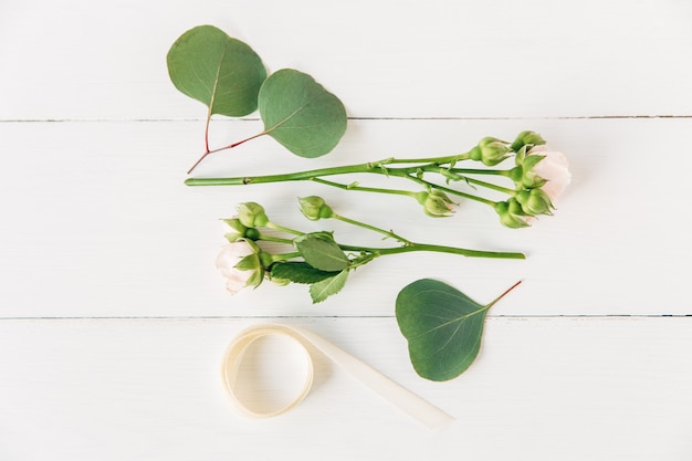 Fresco bouquet di piccole rose rosa con boccioli, foglie verdi, nastro bianco. Superficie piatta in legno minimalista, bianca.