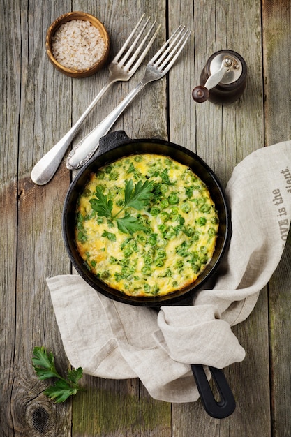 Fritatta con patate, piselli ed erbe aromatiche in una padella di ferro su una vecchia superficie di legno. Messa a fuoco selettiva. Stile rustico.