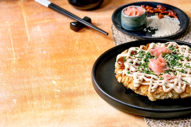 Frittella di cavolo okonomiyaki giapponese fast food fatta in casa decorata da cipollotto, zenzero sottaceto, salsa mayo su piatto in ceramica nera con bacchette e ingredienti sopra. Tabella delle texture.