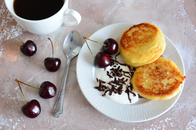 frittelle di formaggio con yogurt, ciliegie e cioccolato su piatto bianco e tazza di caffè