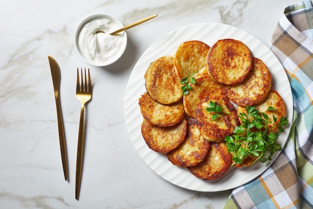 Frittelle di patate con ricotta con aglio, prezzemolo, servite con salsa di panna acida su un piatto su uno sfondo di pietra di marmo chiaro con posate dorate, vista dall'alto, primo piano