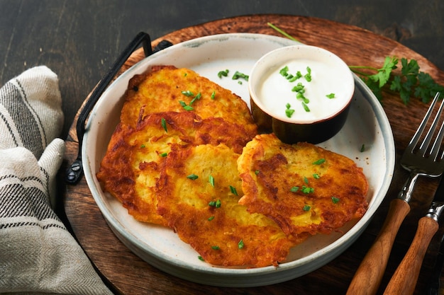 Frittelle di patate Frittelle di patate fritte fatte in casa o latkes con panna e cipolle verdi in un piatto rustico su vecchio sfondo di tavolo in legno nero Stile rustico Cibo sano Vista dall'alto