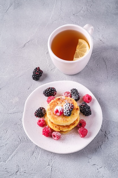 Frittelle di ricotta e zucchero a velo, frittelle di ricotta dessert con lamponi e bacche di more nella piastra vicino alla tazza di tè caldo con fetta di limone