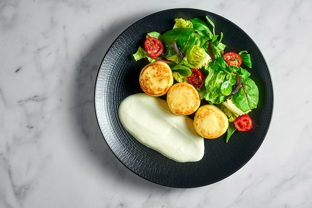 Frittelle di ricotta salate con panna acida, pomodorini al forno e insalata, servite in un piatto nero su una superficie di marmo