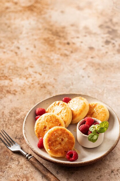 Frittelle di ricotta syrniki con topping al miele e lampone e menta su piatto in ceramica con forchetta su sfondo testurizzato Spazio di testo