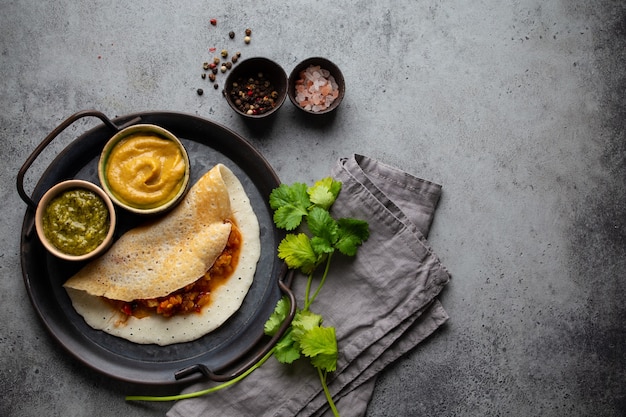 Frittelle di riso indiane tradizionali Dosa con ripieno di sambar di verdure, chutney di salse, su piastra metallica, tavolo di pietra sullo sfondo. Pasto veloce o spuntino vegetariano del sud dell'India, vista dall'alto