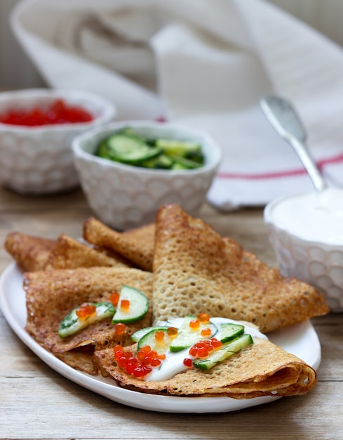 Frittelle di segale e cereali integrali servite con panna acida, caviale e cetrioli. Stile rustico.