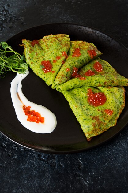 Frittelle di spinaci verdi sottili classiche con panna acida e caviale rosso in un piatto nero.