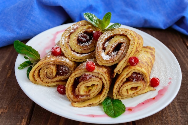 Frittelle dorate sotto forma di rotolo con marmellata di fragole e zucchero a velo su un piatto bianco su un tavolo di legno. Avvicinamento. Colazione