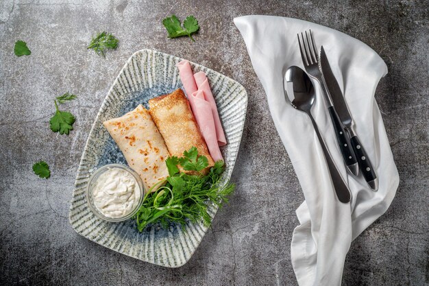 Frittelle ripiene di prosciutto in un piatto con prezzemolo e aneto e panna acida su una tavola di pietra grigia, flatlay