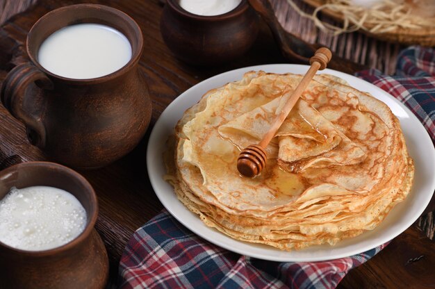 Frittelle sottili fatte in casa con miele impilate in una pila, su un tavolo di legno con una tazza di latte, una pentola di panna acida e uova in un cestino. Slavonia tradizionale, festa pagana (Maslinitsa). Stile country f