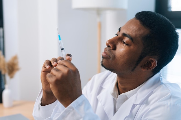 Fronte del primo piano del medico maschio nero pensieroso in uniforme bianca premurosa che osserva alla siringa con il vaccino in mano che si siede alla scrivania nella clinica medica