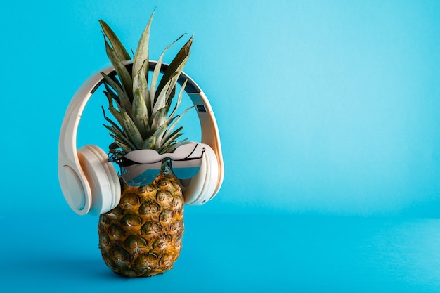 Fronte divertente creativo dell'ananas che indossa le cuffie degli occhiali da sole Faccia di ananas che levita ascoltando musica su sfondo estivo di colore blu con spazio di copia. Foto d'archivio di alta qualità.