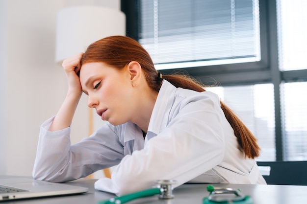Fronte ravvicinato di una giovane dottoressa stanca esausta in camice bianco seduta alla scrivania con laptop e guardando lo schermo.