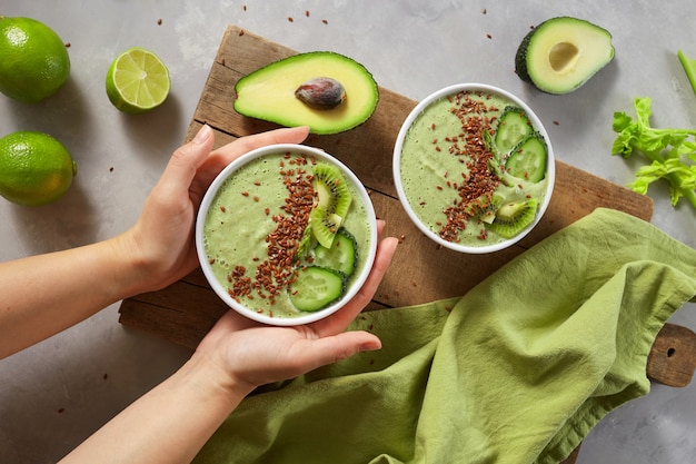 Frullato di avocado e cetriolo fresco. Mani femminili che tengono un piatto di frullati di verdure con kiwi e semi di lino contro una tavola di legno. Vista dall'alto