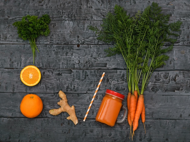 Frullato di carota appena fatto, mazzo di carote, arancia e radice di zenzero su un rustico tavolo scuro.