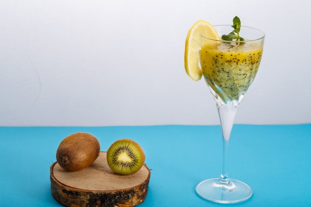 Frullato di kiwi guarnito con menta e limone in un bicchiere su una superficie blu vicino al kiwi