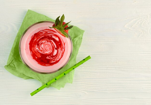 Frullato di latte alla fragola con sciroppo in una vista dall'alto di vetro. Cocktail di proteine con fragole e sciroppo su carta verde. Sfondo rustico in legno bianco. Copia spazio. Bicchiere, bacche, cannuccia di carta