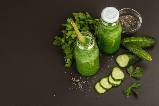 Frullato verde con cetriolo in bottiglie di vetro. Verdure fresche mature, verdure e semi di chia. Sfondo di cemento in pietra nera, vista dall'alto