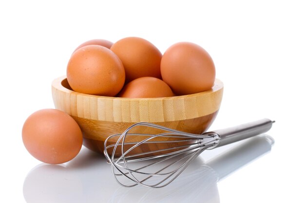 Frusta in metallo per montare uova e uova marroni in una ciotola di legno isolata su bianco