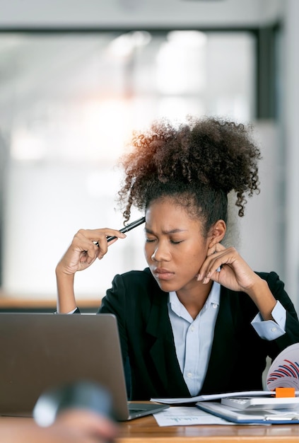 Frustrata giovane donna afroamericana che tiene gli occhi chiusi dopo aver lavorato a lungo al computer portatile in ufficio sentendosi stanca e stressata