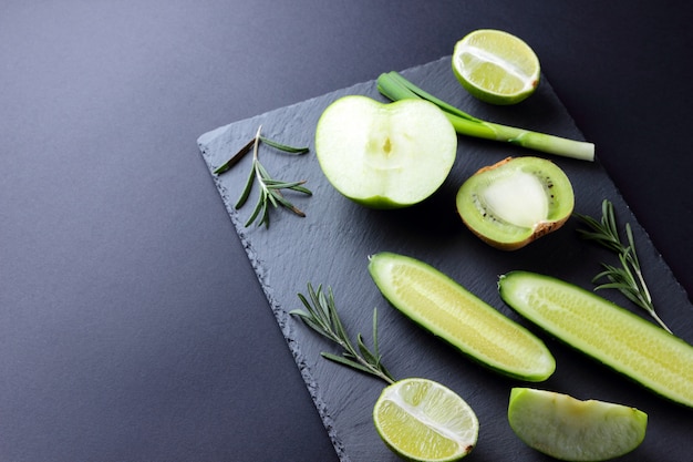 Frutta e verdura verdi sul bordo scuro dell'ardesia. Concetto di prodotti verdi naturali. Avocado, kiwi, lime e mela. Rosmarino, aneto ed erba cipollina su tavola di pietra