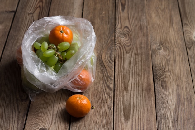 Frutta (mandarini, mele, uva) in un sacchetto di plastica su un tavolo di legno naturale. L'immagine mostra gli effetti dannosi dei sacchetti di plastica sul cibo.