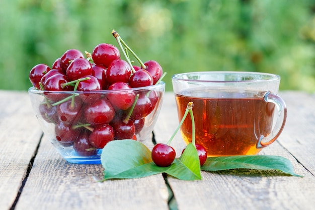 Frutti di ciliegia rossa con peduncoli in ciotola di vetro e tazza di tè su vecchie tavole di legno