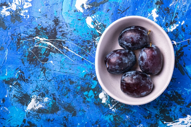 Frutti di prugna dolce freschi in ciotola bianca su fondo astratto blu, spazio della copia di vista superiore