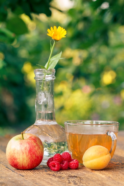 Frutti maturi di mela, albicocca e fragole, fiore di calendula con gambo in una boccetta di vetro e tazza di tè verde su tavole di legno con sfondo naturale sfocato