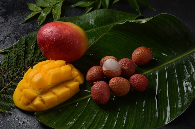 Frutto di mango fresco con pezzi di mango a dadini a fette su un piatto e litchi freschi e sbucciati che mostrano la pelle rossa e la polpa bianca su sfondo scuro.