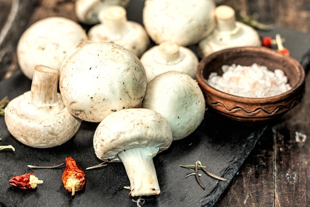 Funghi champignon freschi con spezie ed erbe aromatiche. Ingrediente crudo per cucinare cibi vegani. Vecchio fondo di legno, luce dura di modo, ombra scura, stile rustico, primo piano