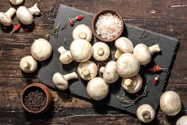 Funghi champignon freschi con spezie ed erbe aromatiche. Ingrediente crudo per cucinare cibi vegani. Vecchio fondo di legno, luce dura di modo, ombra scura, stile rustico, vista dall'alto