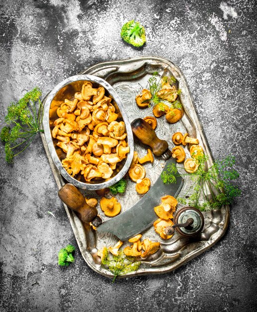 Funghi finferli freschi con erbe aromatiche e un vecchio coltello su un vassoio in acciaio su un tavolo rustico.