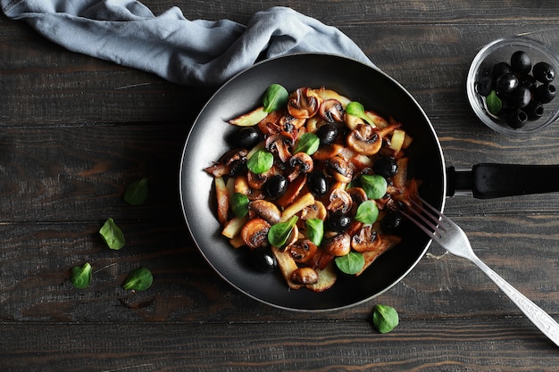 Funghi fritti con le patate, le olive e le verdure in una padella su una vista superiore dell'alimento vegetariano della tavola di legno.