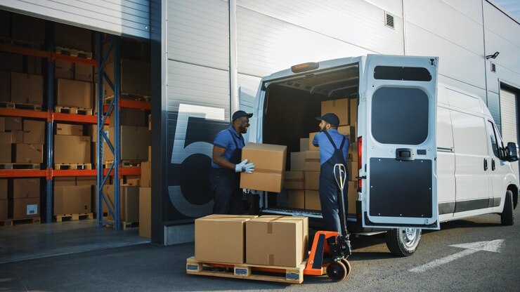 Foto fuori del magazzino di distribuzione logistica due lavoratori parlano scherzo uso camion a palette a mano inizio carico camion di consegna con scatole di cartone ordini online acquisti beni di commercio elettronico