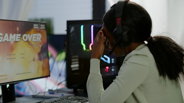 Furiosa giocatrice africana con auricolare che perde il videogioco sparatutto spaziale con grafica durante il campionato di gioco giocando da casa al potente personal computer RGB, esibendosi al torneo di eSports