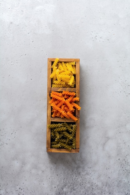 Fusilli crudi multicolori tre colori sparsi in una scatola di legno su una superficie di cemento grigio. Vista dall'alto.