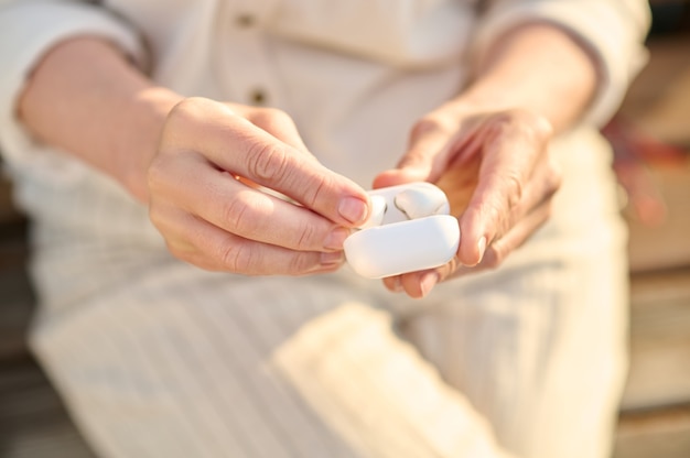 Gadget moderno. Donna in abiti leggeri con cuffie wireless in una piccola custodia bianca all'aperto, il viso non è visibile