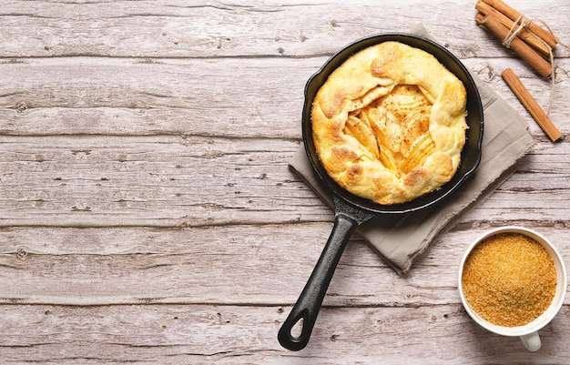 Galette di mele in una padella nera zucchero di canna in una tazza e bastoncini di cannella su un tavolo di legno