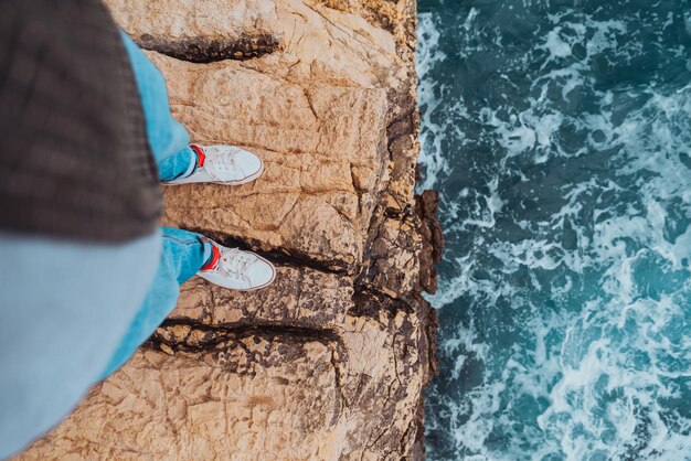 Gambe dell'uomo in jeans e scarpe da ginnastica bianche sul bordo della scogliera vista del mare in tempesta blu