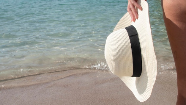 Gambe di donna con un cappello bianco in mano vicino all'acqua cristallina del mare